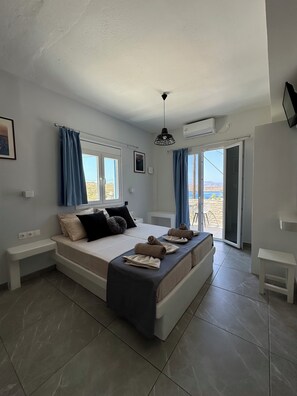 A bedroom with a large bed, a ceiling fan, an air conditioning unit, a window with curtains, and a view of the outdoors. - Princess Sissy Hotel (Ios)