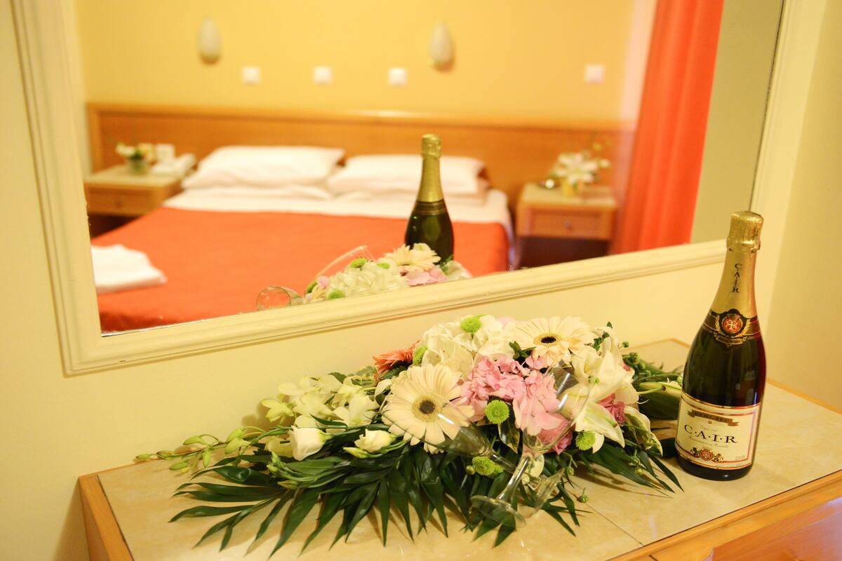 Una habitación de hotel con una cama, una mesita de noche, una botella de champán y un arreglo floral sobre la mesita.