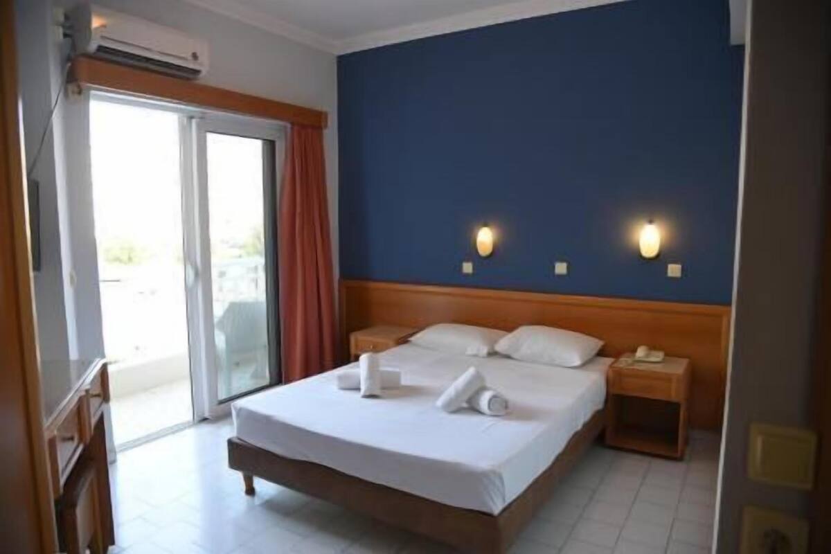 Habitación de hotel con cama, dos mesitas de noche, puerta corredera de cristal y una pared azul decorativa.