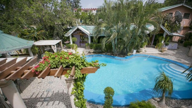 A swimming pool surrounded by lush greenery and potted plants, with a pergola and a small gazebo nearby. - Palm Inn Hotel (Port-au-Prince)
