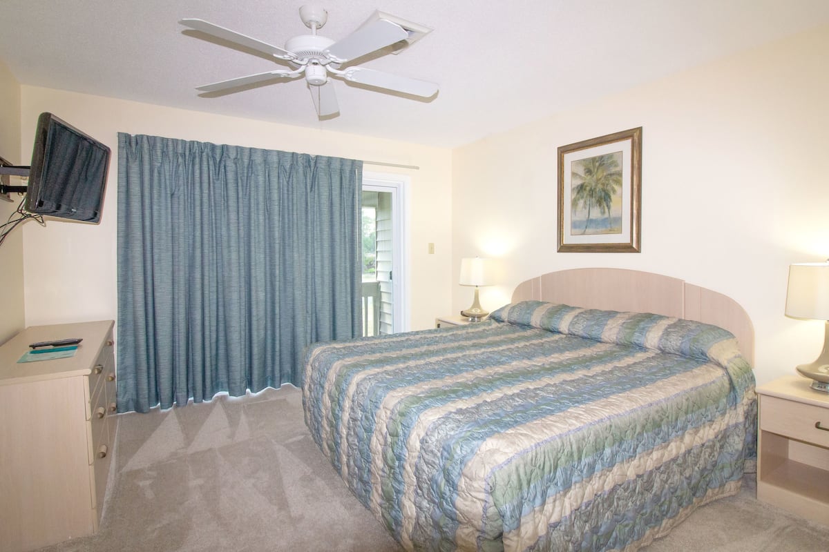 A bedroom with a bed, a ceiling fan, a TV mounted on the wall, a desk, and a nightstand with a lamp.