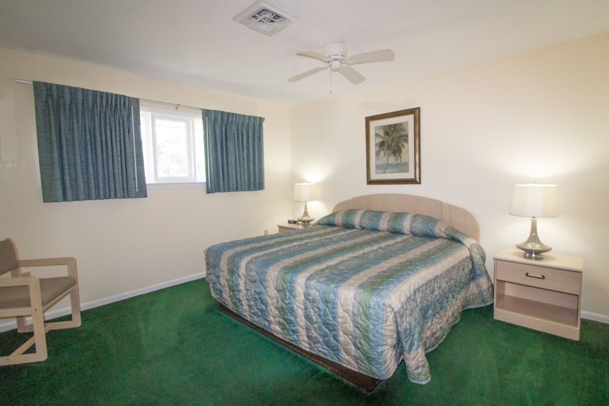 A bedroom with a bed, a chair, a nightstand with a lamp, a ceiling fan, and a window with curtains.