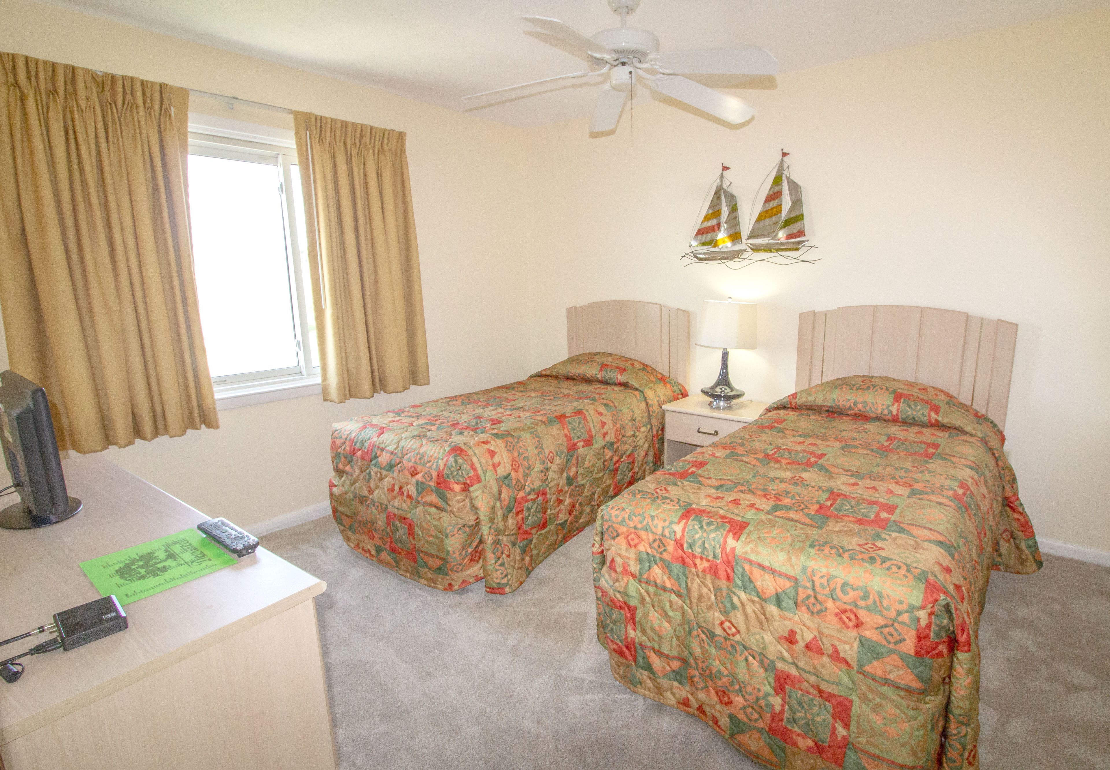 A bedroom with two beds, a desk, a ceiling fan, and a window with curtains.