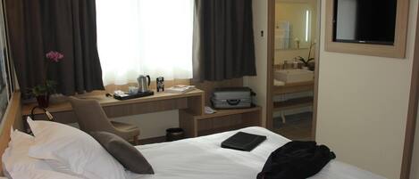 A hotel room with a bed, a desk, a chair, a TV, and a bathroom mirror.