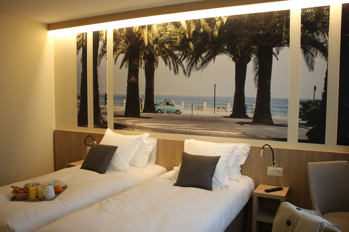 A hotel room with two beds, a nightstand, a chair, and a large beach view mural.