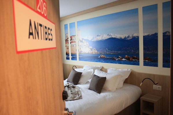 A hotel room with a bed, pillows, a nightstand, and a wall with a scenic view. - Brit Hotel Montargis (Mormant-sur-Vernisson)