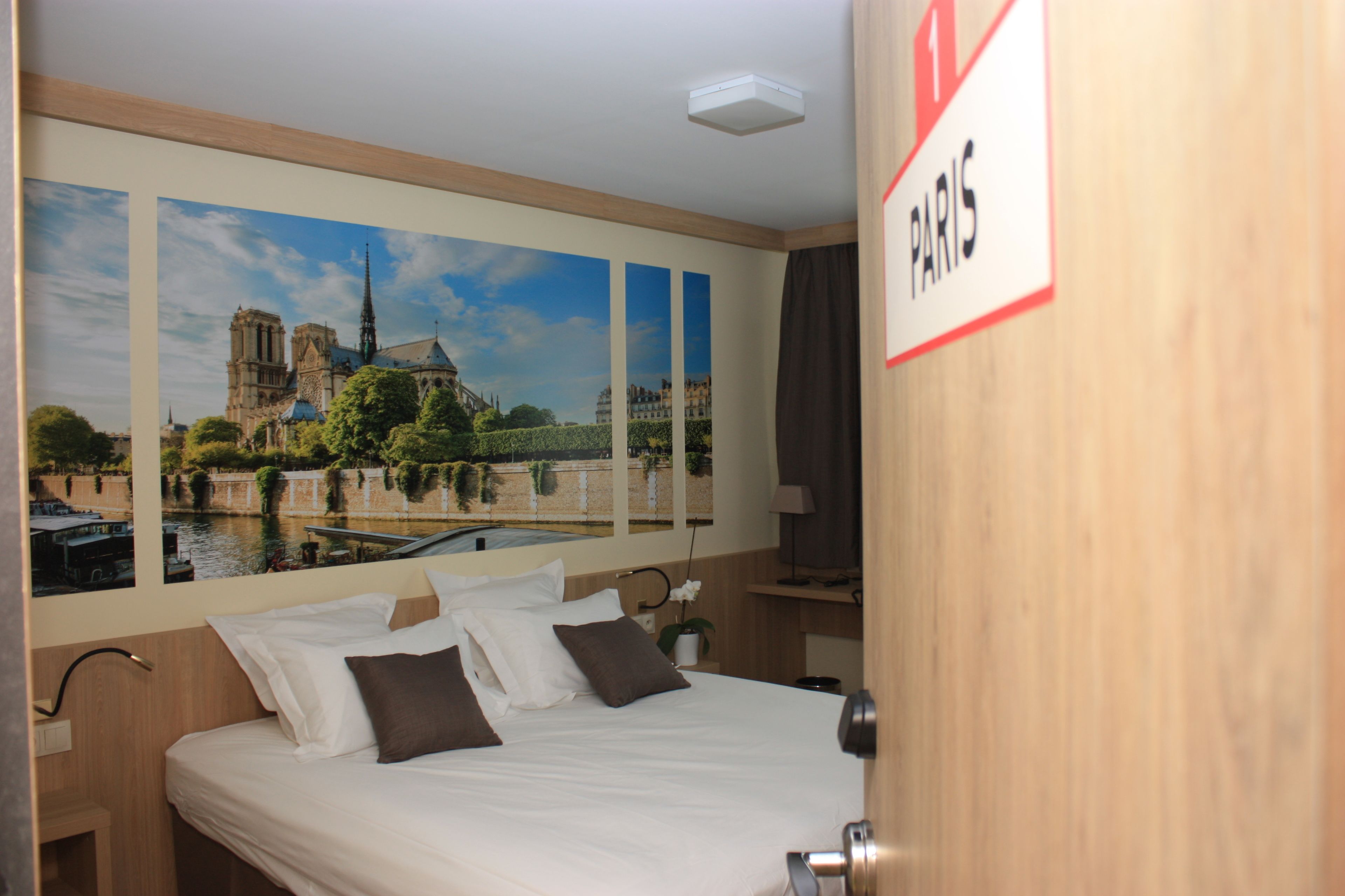 A hotel room with a bed, a nightstand, a lamp, and a wall-mounted sign indicating the location as Paris.