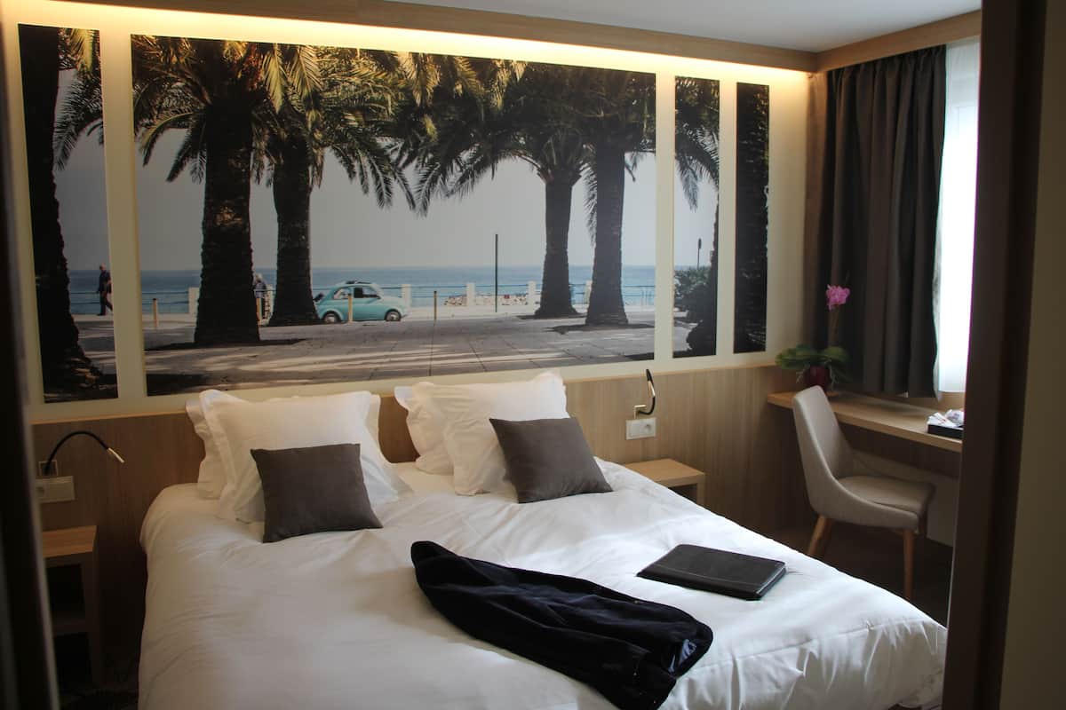 A hotel room with a bed, a desk, a chair, and a large window with a beach view.