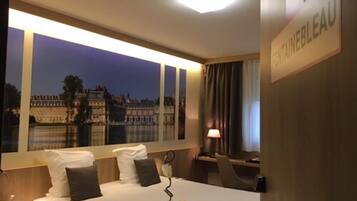 A hotel room with a bed, a nightstand, a lamp, and a view of a cityscape through a large window.
