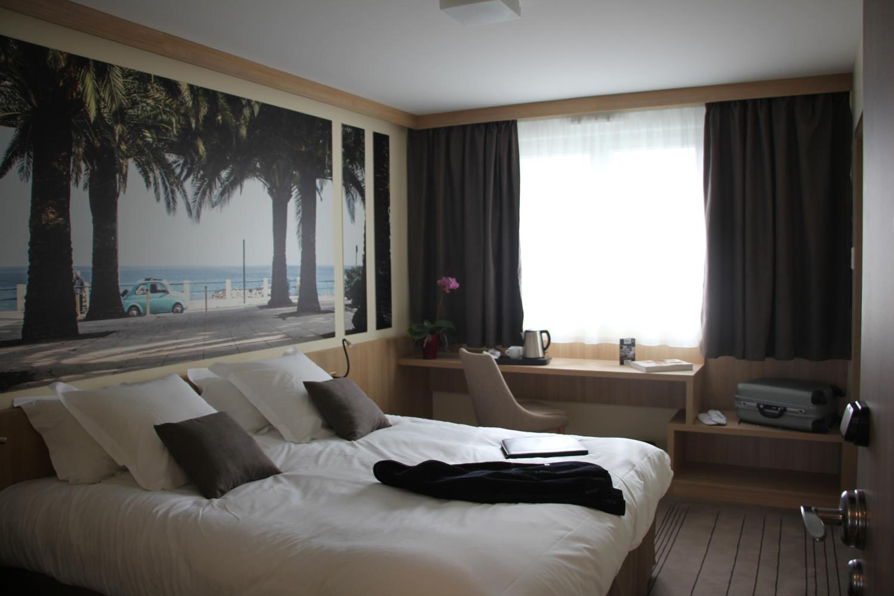 Une chambre d’hôtel avec un lit, un bureau, une chaise, une fenêtre avec des rideaux et une vue sur des palmiers et une voiture sur la plage.