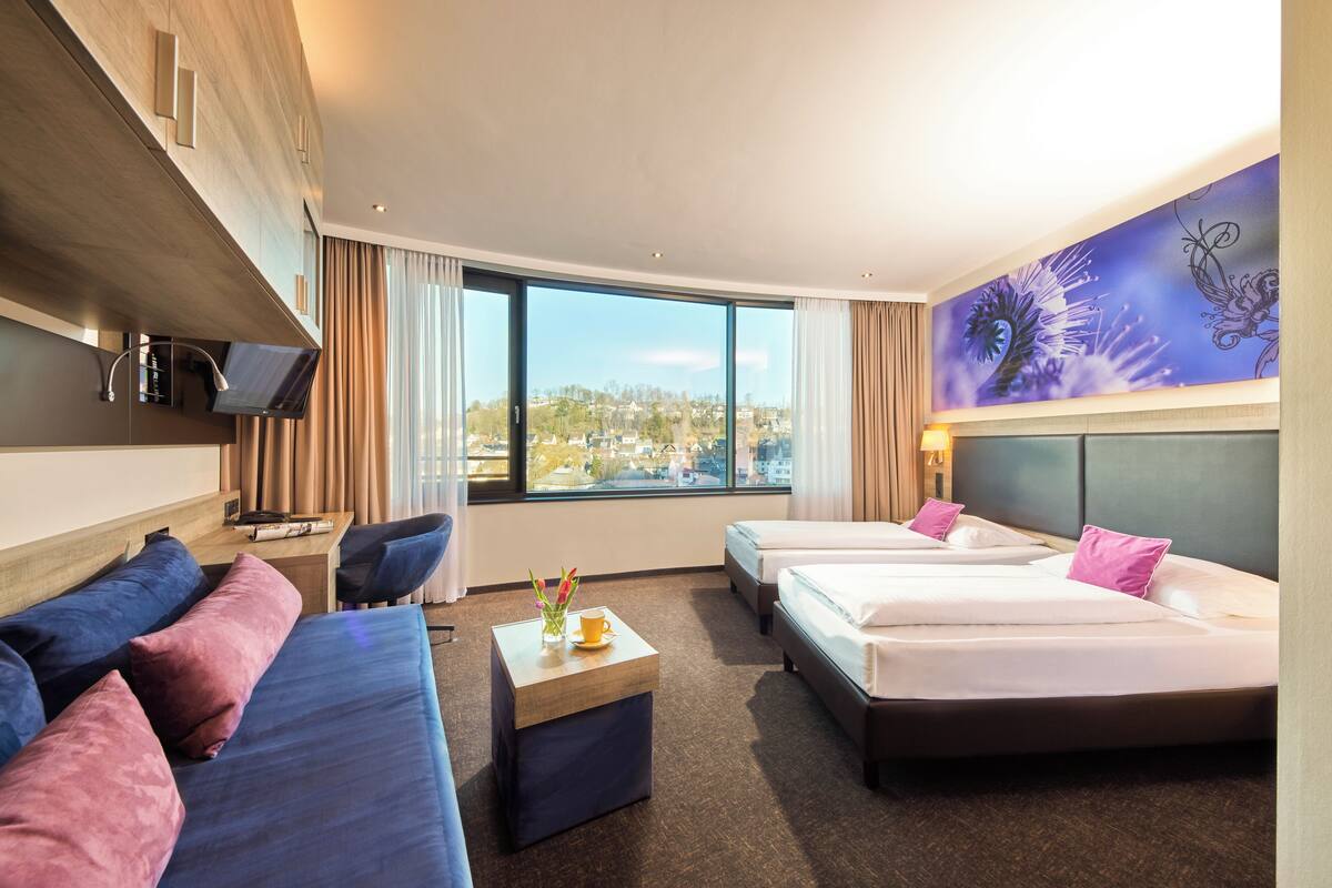 A hotel room with two beds, a sofa, a small table with a cup and flowers, and a large window offering a city view.