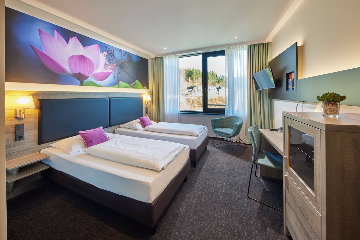 A hotel room with two beds, a desk, a chair, a TV, and a large window with curtains.