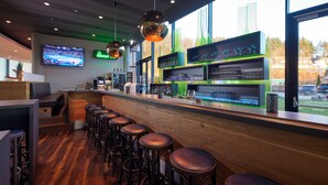 A bar area with wooden flooring, bar stools, a counter with shelves of glasses and bottles, and a TV mounted on the wall. - HANSE HOTEL Attendorn (Attendorn)