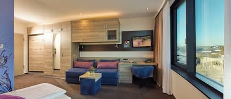 A modern hotel room with a kitchenette, a sofa, a small table, and a large window offering a city view.