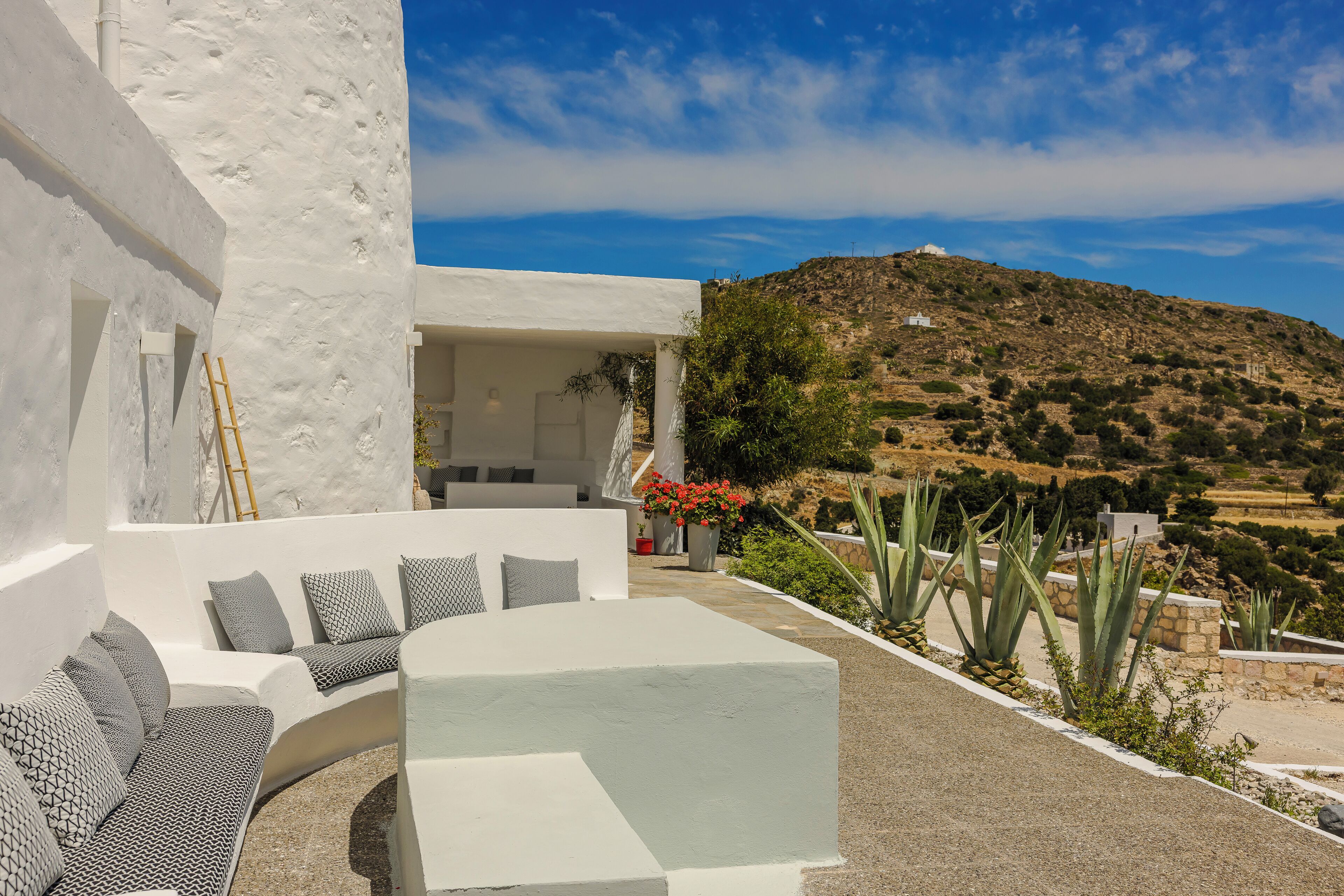 A modern outdoor seating area with white walls and grey patterned cushions, surrounded by a landscaped garden with agave plants and a view of a hilly landscape.