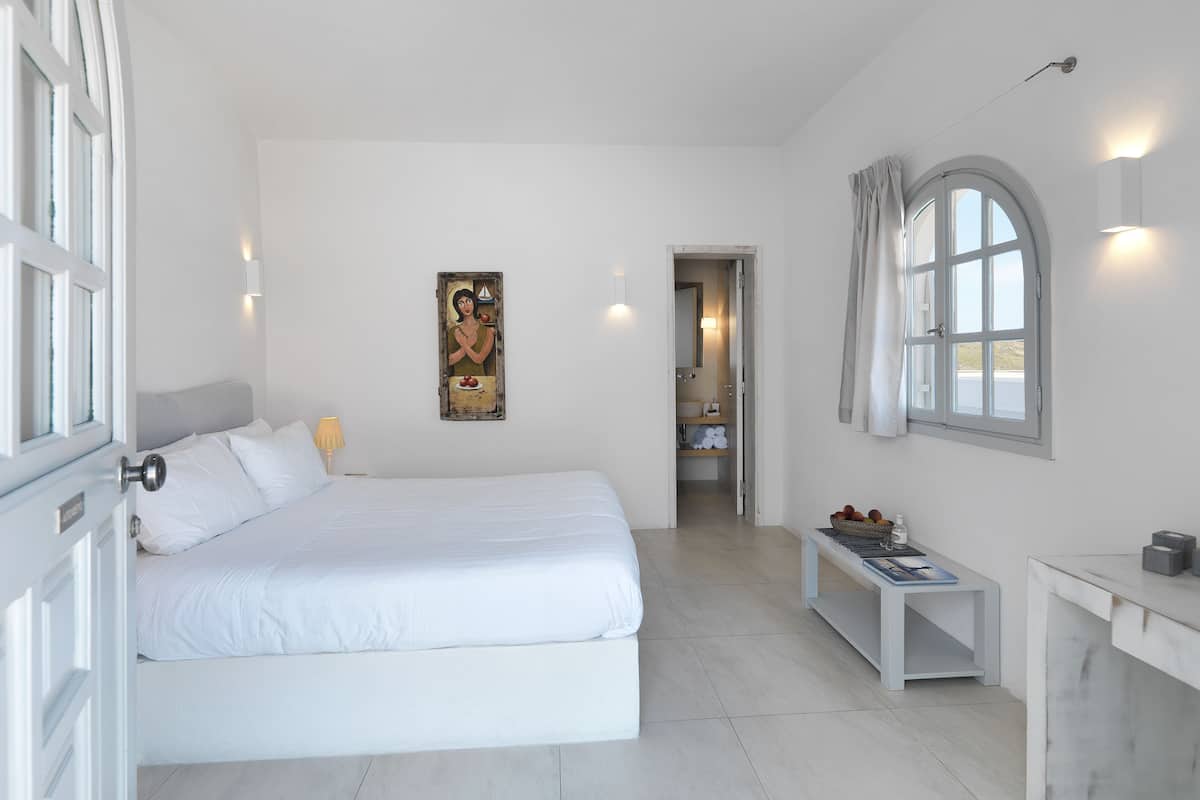 A minimalist bedroom with a large bed, a bedside table, a small desk, and a window with curtains.