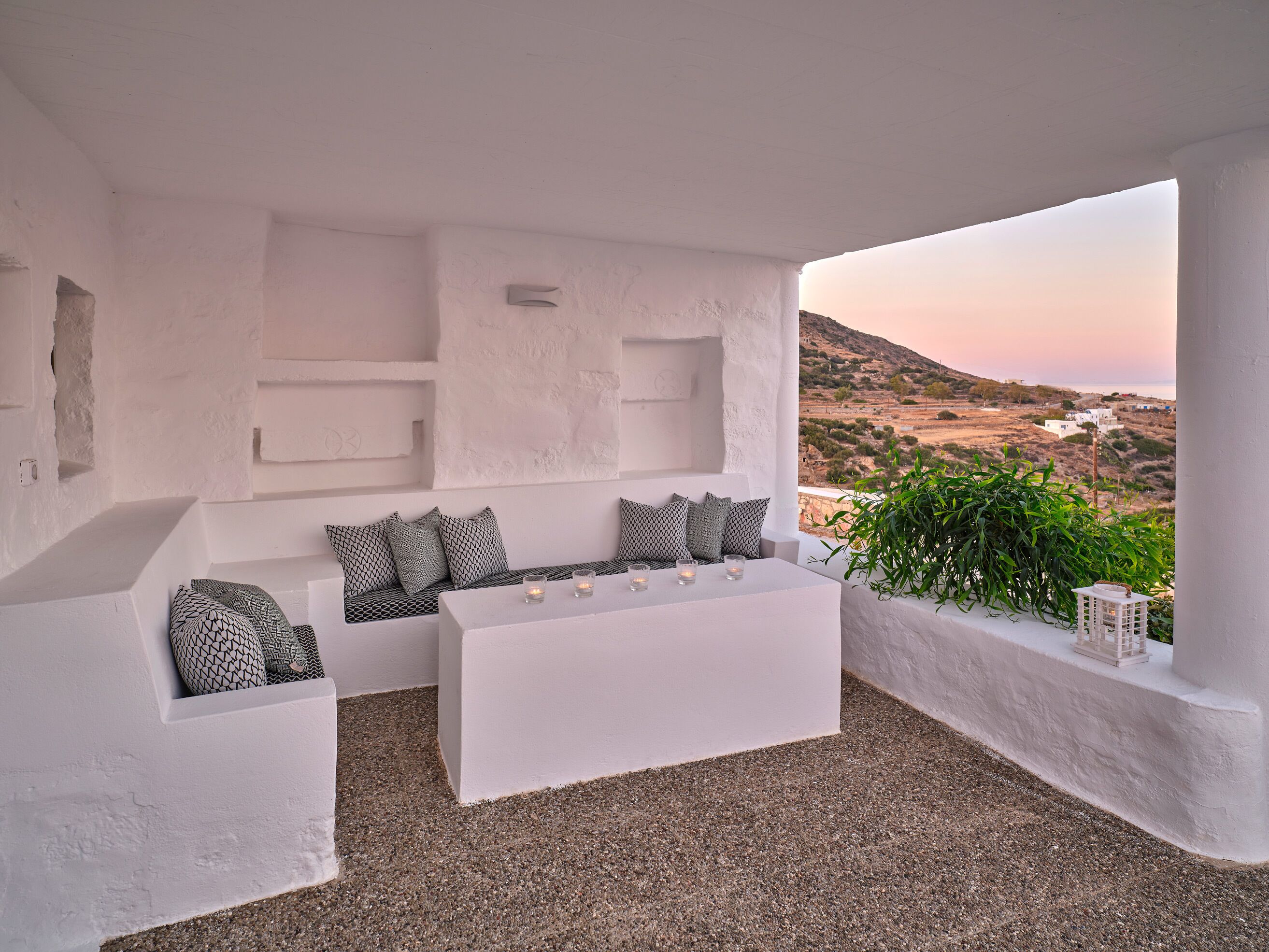 A white, open-plan living area with a sofa and coffee table, a view of a hilly landscape, and a small potted plant.