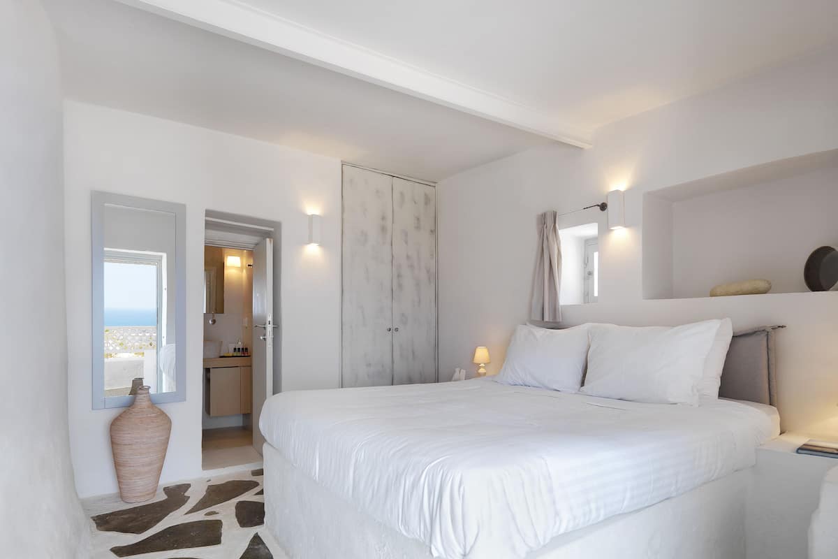 A bedroom with a large bed, a mirror, a nightstand, and a view of the sea through an open door.