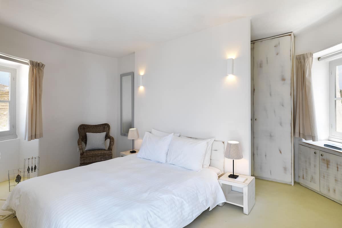 A bedroom with a large bed, two bedside tables with lamps, a chair, a wardrobe, and a window with curtains.