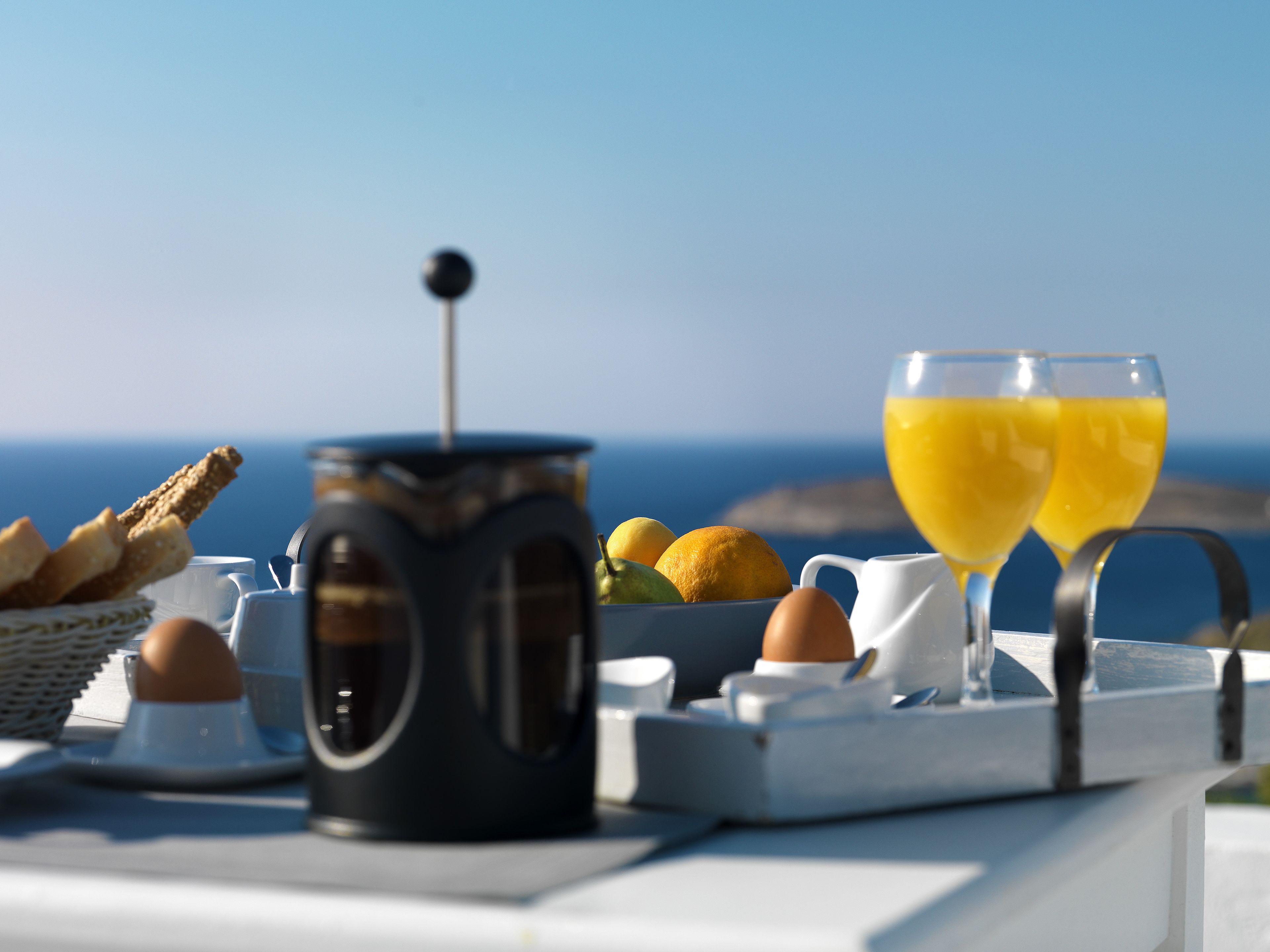 A breakfast setup with a coffee pot, eggs, toast, and orange juice glasses on a table with a scenic ocean view in the background.