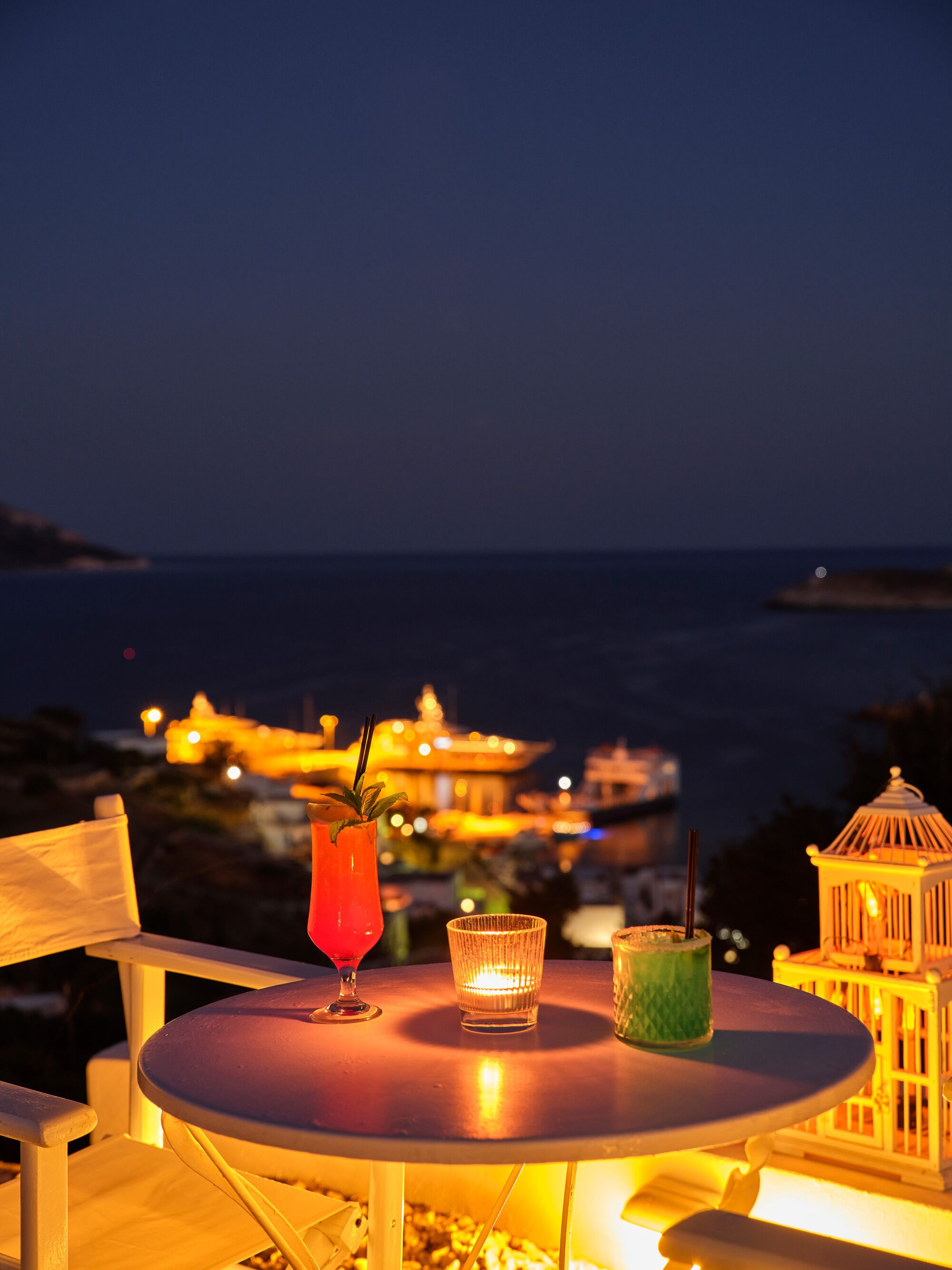 A table with drinks and a candle, overlooking a coastal view with buildings and lights.