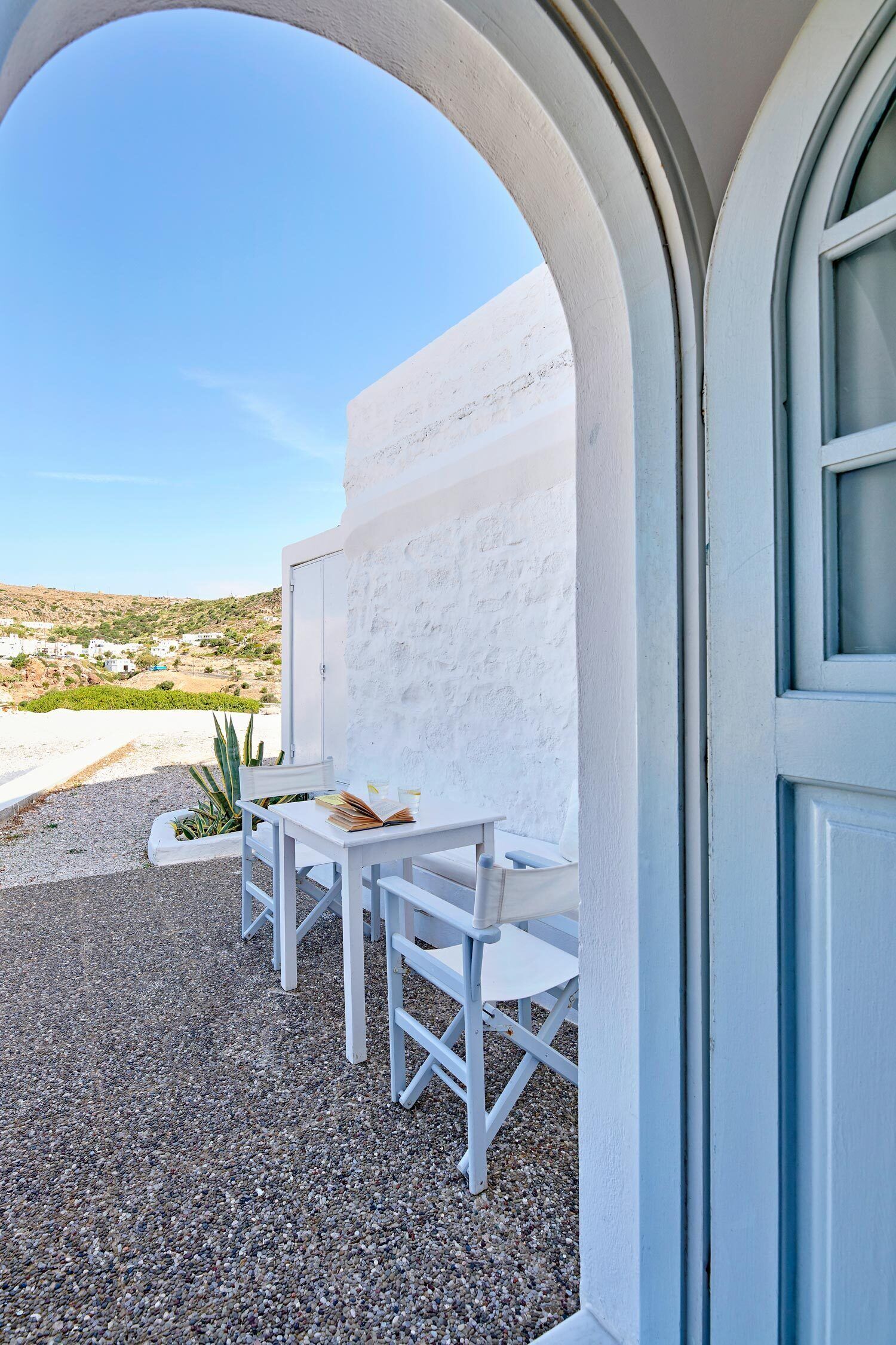 Un espace extérieur aménagé avec des meubles blancs, comprenant une table et des chaises, un mur blanc et une vue sur une colline.