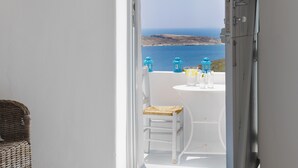A room with a bed, a wicker chair, and a table with two glasses and a bottle. A door leads to a balcony with a view of the sea and two blue lanterns. - The Windmill Kimolos (Kimolos)