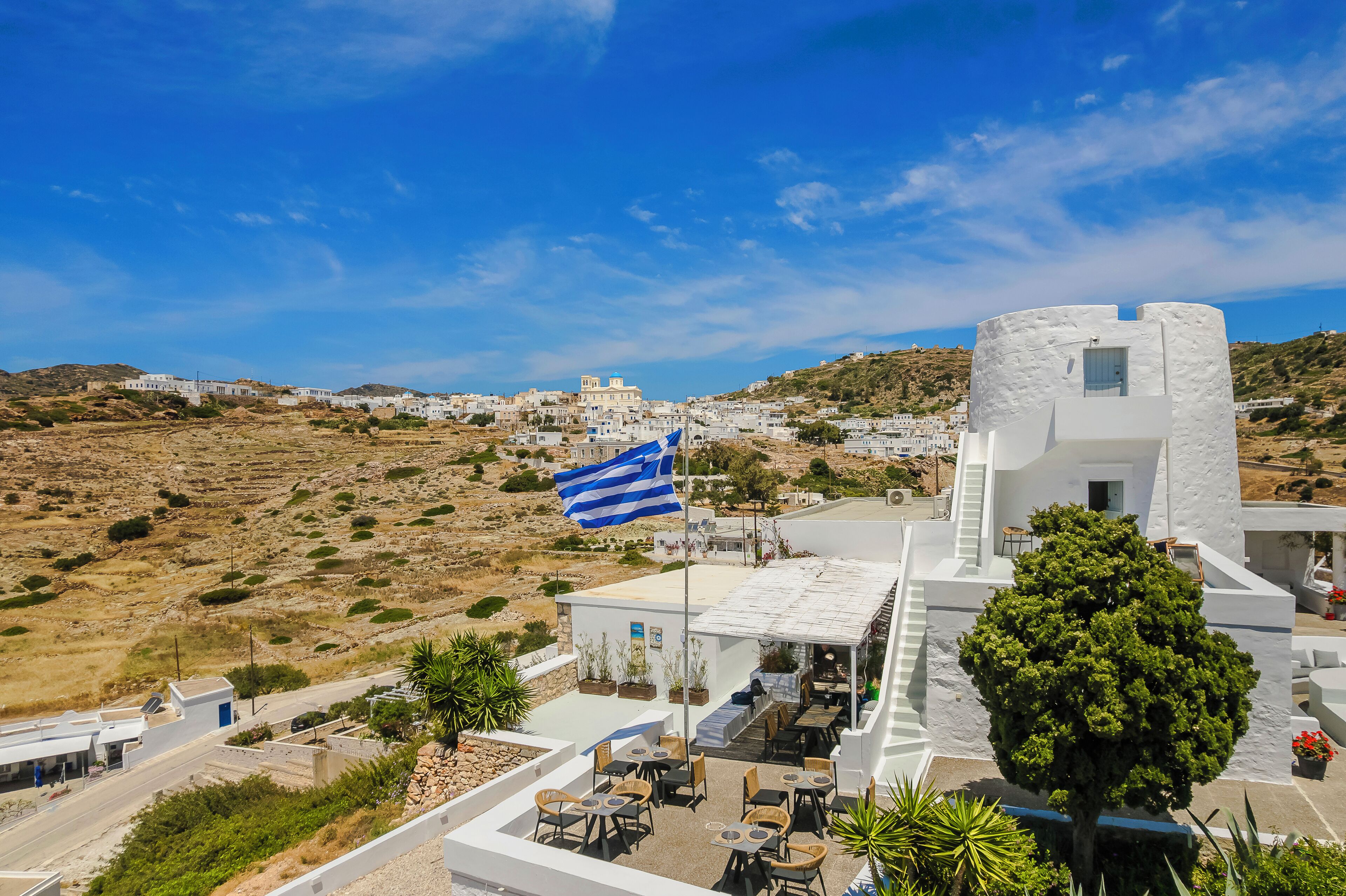 Un pueblo costero con edificios blancos, una bandera y mesas al aire libre.