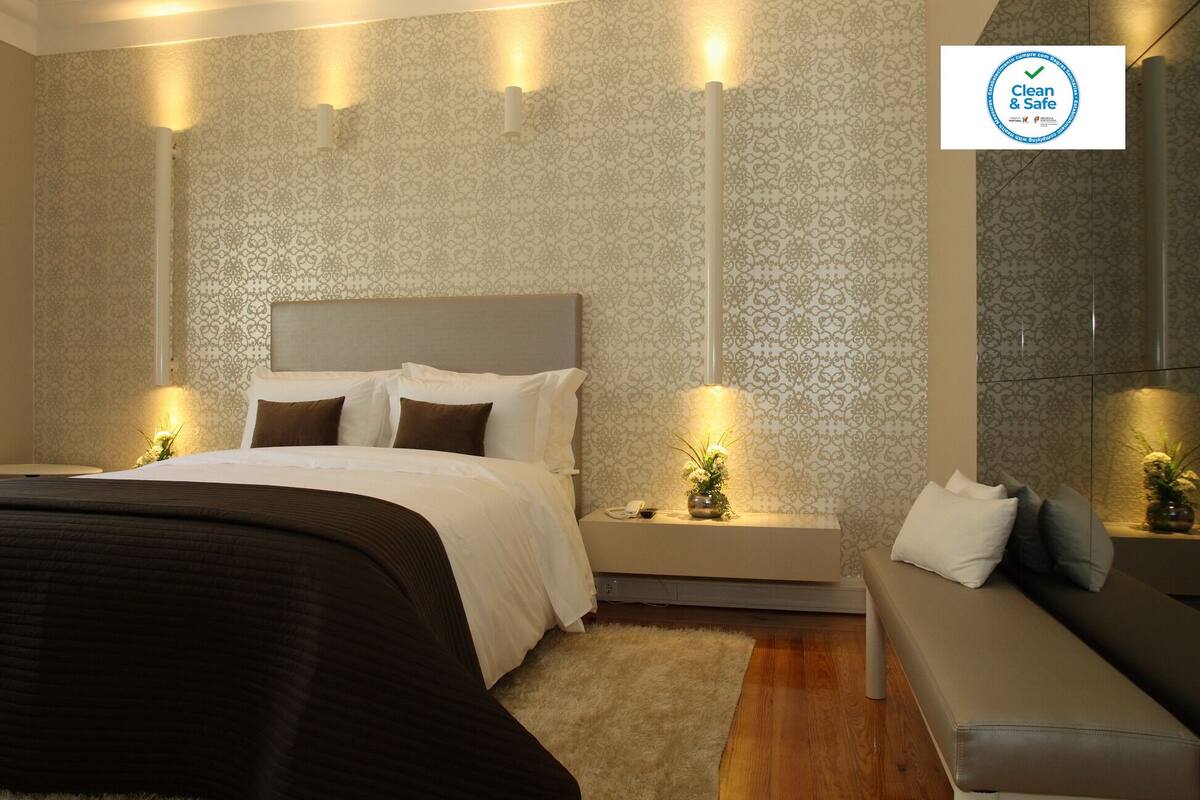 A modern hotel room with a large bed, bedside tables with lamps, a sofa, and a patterned wallpaper.
