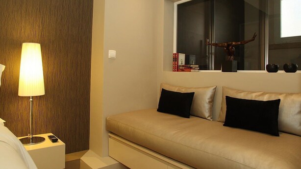 A modern hotel room with a beige sofa, a lamp, a remote control, and a window with a decorative item. - Castilho House (Lisbon)