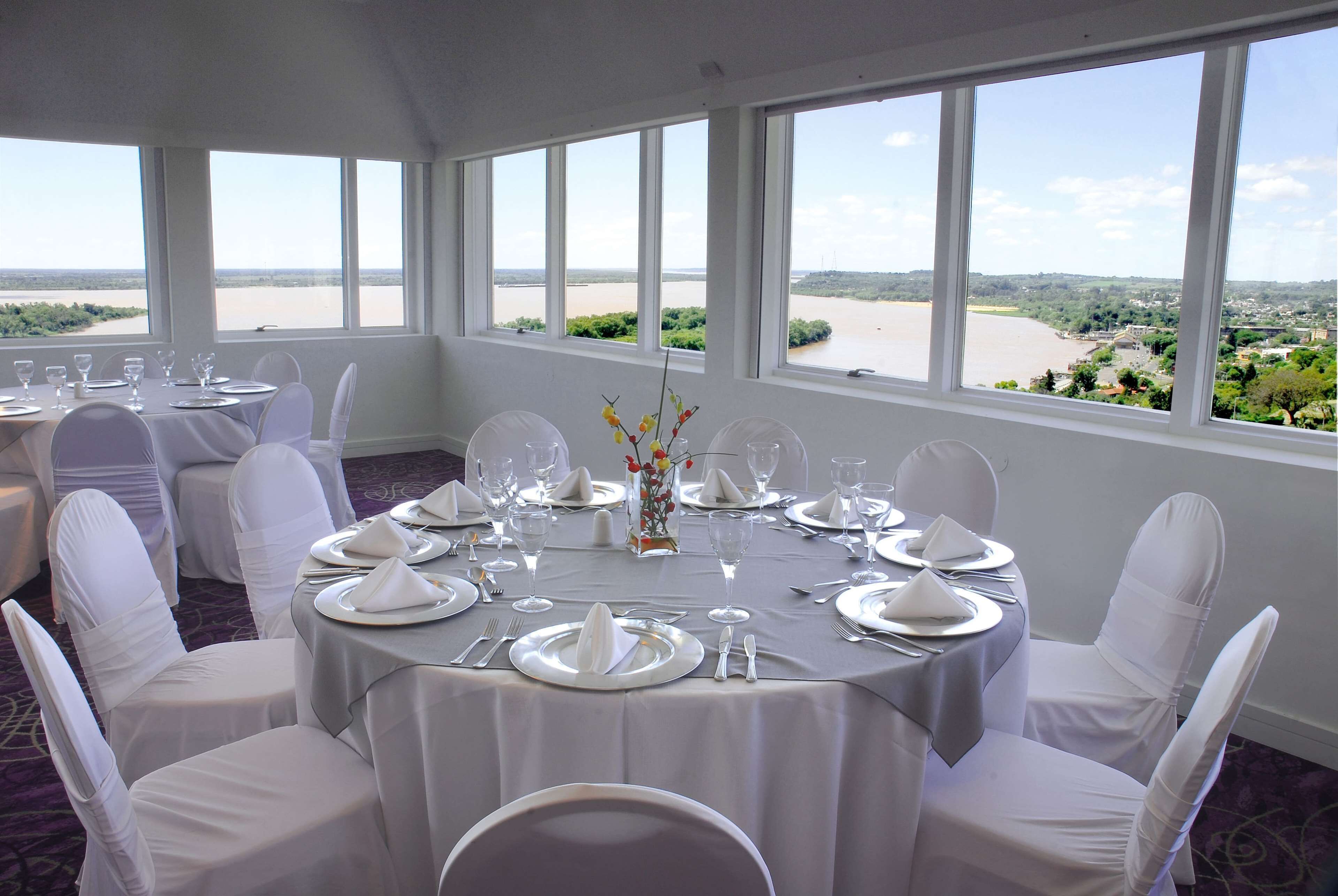 Un salón de banquetes con mesas redondas dispuestas para un evento formal, amplias ventanas que ofrecen vistas a un río y follaje.