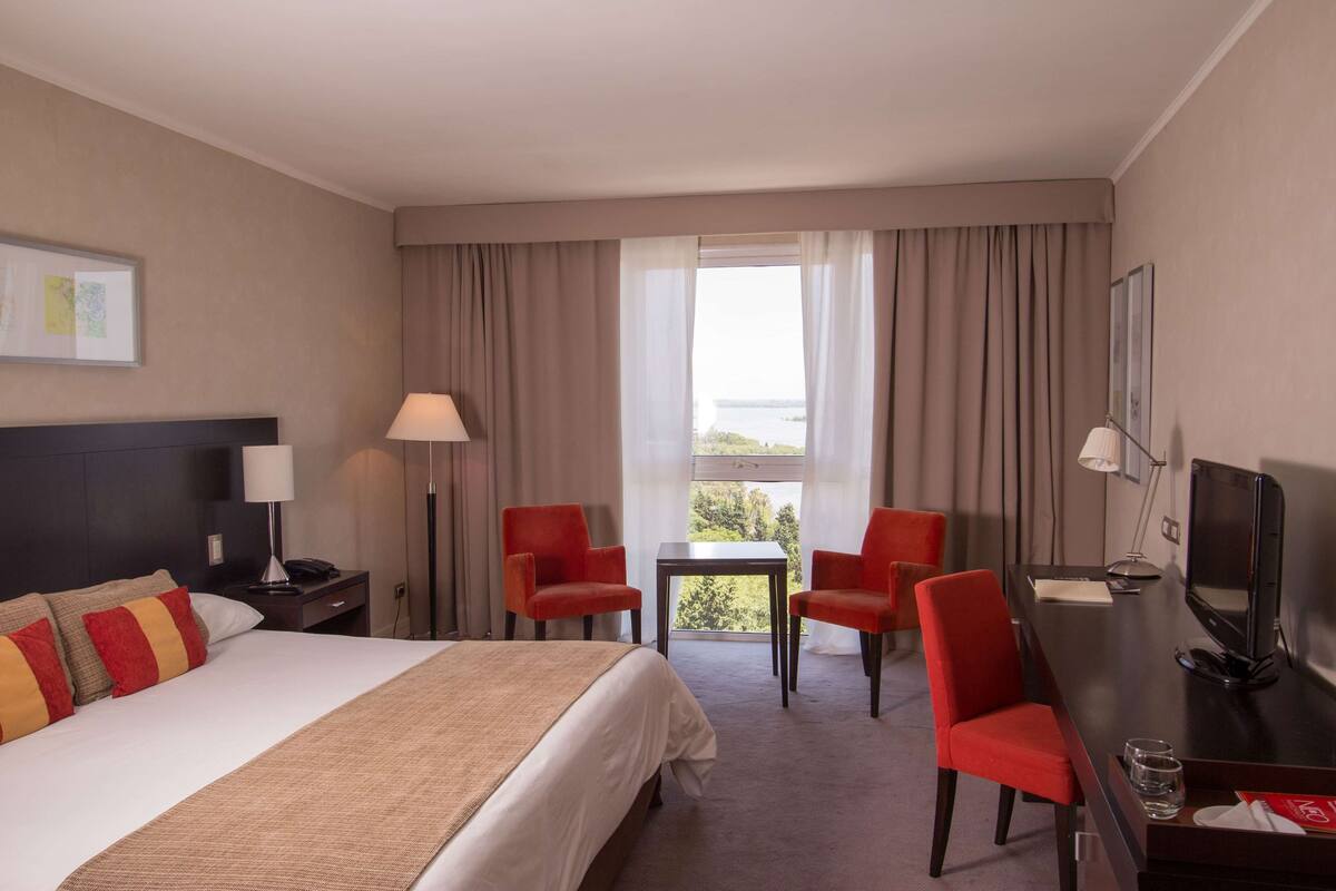 A hotel room with a bed, two red chairs, a desk with a TV, a lamp, and a view of the outdoors.