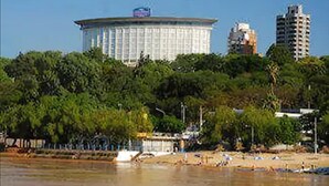 Un edificio circular grande con una cúpula, rodeado de abundante vegetación y una playa con gente y barcos.