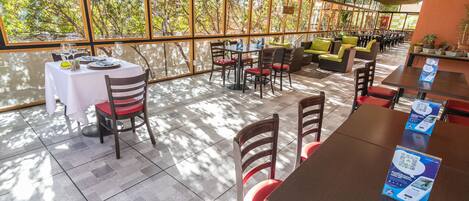 Un vaste espace extérieur aménagé pour les repas, avec des tables et des chaises, de grandes fenêtres offrant une vue sur les arbres et un toit de chaume.