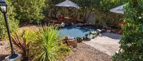 A backyard with a swimming pool, surrounded by rocks and plants, and two umbrellas providing shade.