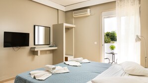 A hotel room with a bed, a TV, a mirror, a small table with a plant, and a balcony with a view of greenery.