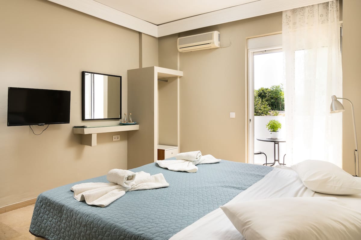 Ein Hotelzimmer mit Bett, Fernseher, Spiegel, einem kleinen Tisch mit Pflanze und einem Balkon mit Blick auf Grünfläche.