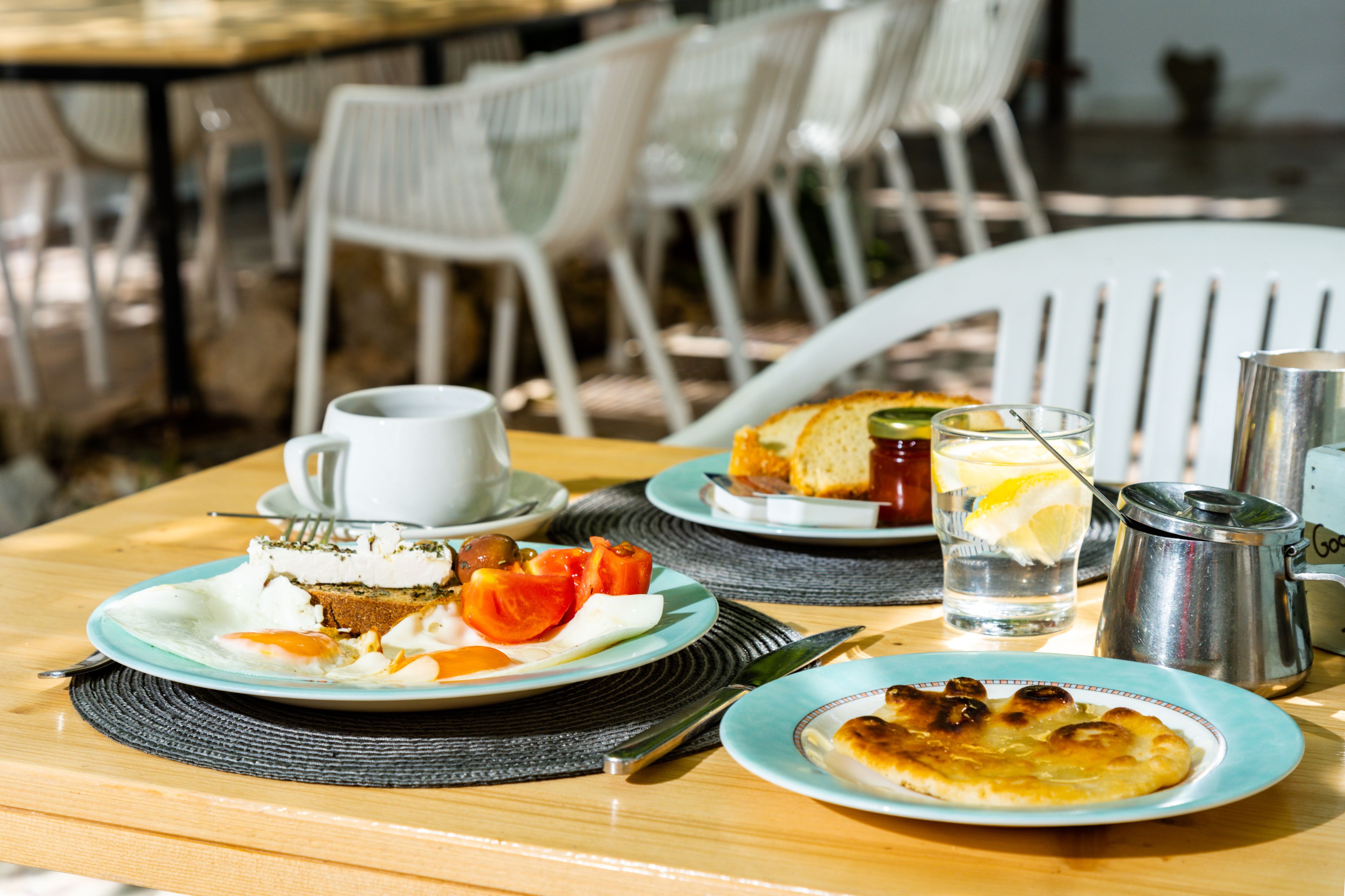Una mesa con alimentos para el desayuno, como tostadas, huevos y una bebida, ubicada al aire libre con sillas al fondo.