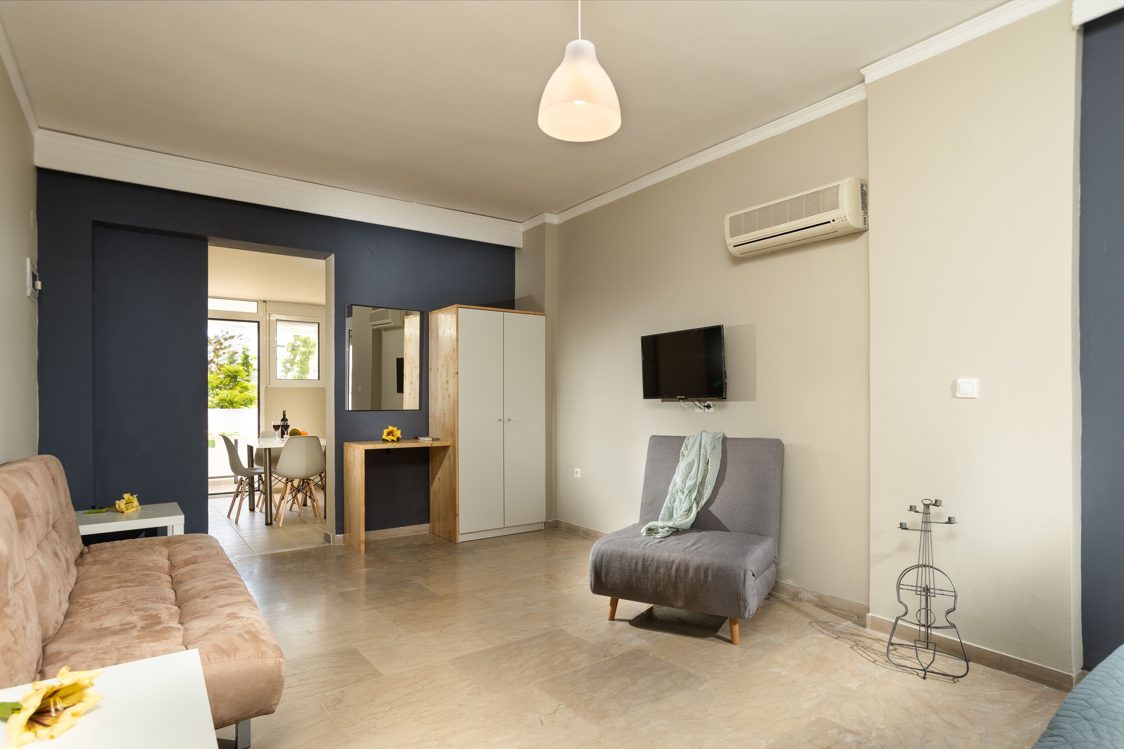 A modern living room with a sofa, a grey armchair, a dining area with a table and chairs, and a TV mounted on the wall.