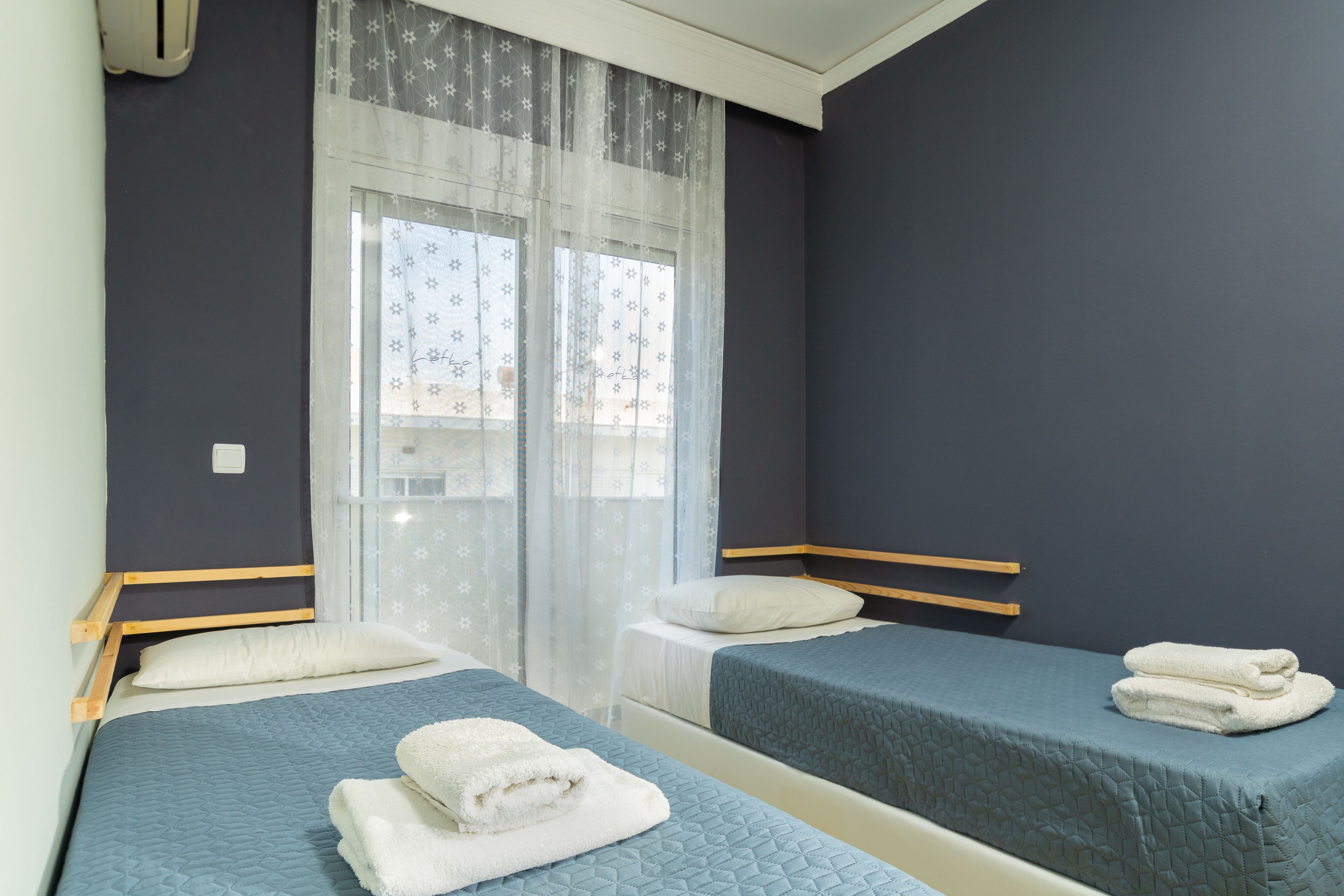 Una habitación con dos camas, cada una con colcha azul acolchada y almohadas blancas. Se ve una ventana con cortinas traslúcidas y un diseño estampado.
