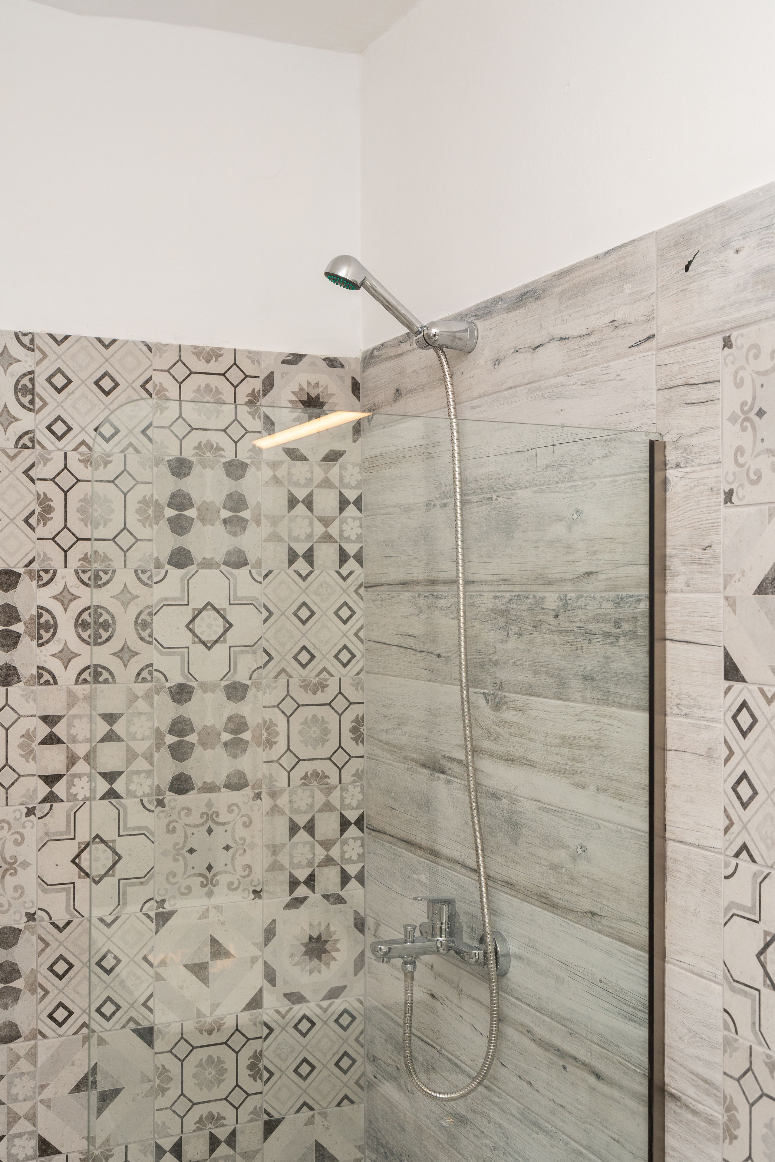 A shower with a handheld showerhead and a wall-mounted faucet, surrounded by patterned tiles.