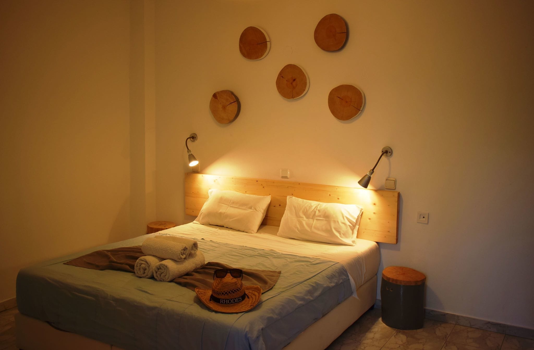 A neatly made bed with a wooden headboard, multiple pillows, and a blanket. There are wall-mounted clocks and a small table with a lamp beside the bed.
