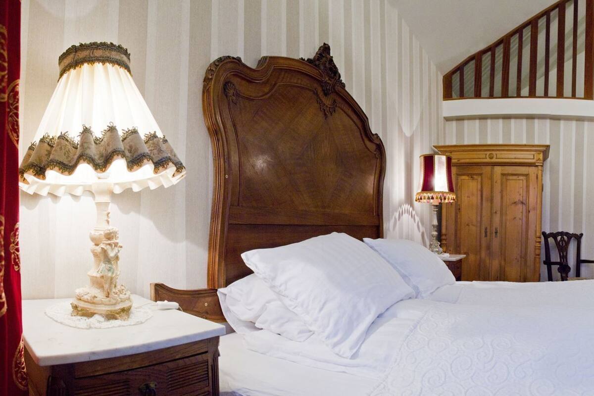 A bedroom with a large bed, a wooden headboard, a nightstand with a lamp, a wooden wardrobe, a chair, and a staircase leading to an upper level.