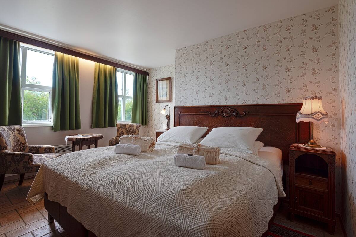 A neatly made bed with a wooden headboard, a bedside table with a lamp, and a chair with a patterned cushion.