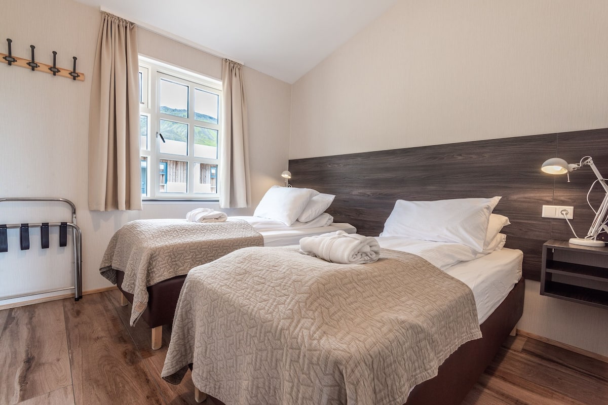 A hotel room with two beds, a window with curtains, a bedside table with a lamp, and a wall-mounted coat rack.