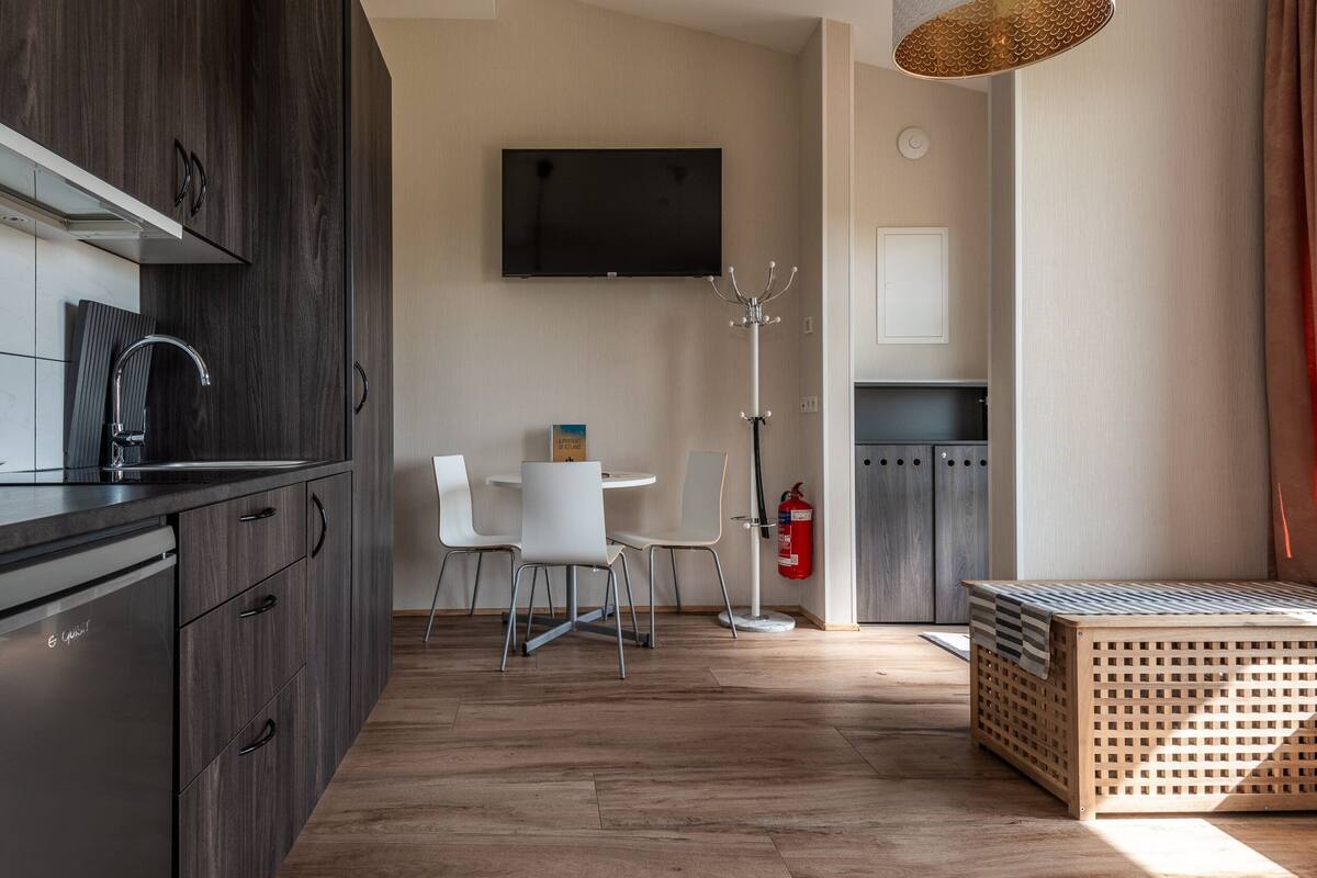 A modern kitchen with dark wood cabinets, a built-in dishwasher, and a flat-screen TV mounted above the counter. There is a dining area with a white table and chairs, and a wooden bench with a striped cushion. A fire extinguisher is mounted on the wall near the dining area.