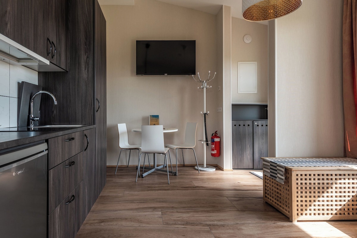 A modern kitchen with dark wood cabinets, a built-in dishwasher, and a flat-screen TV mounted above the counter. There is a dining area with a white table and chairs, and a wooden bench with a striped cushion. A fire extinguisher is mounted on the wall near the dining area.