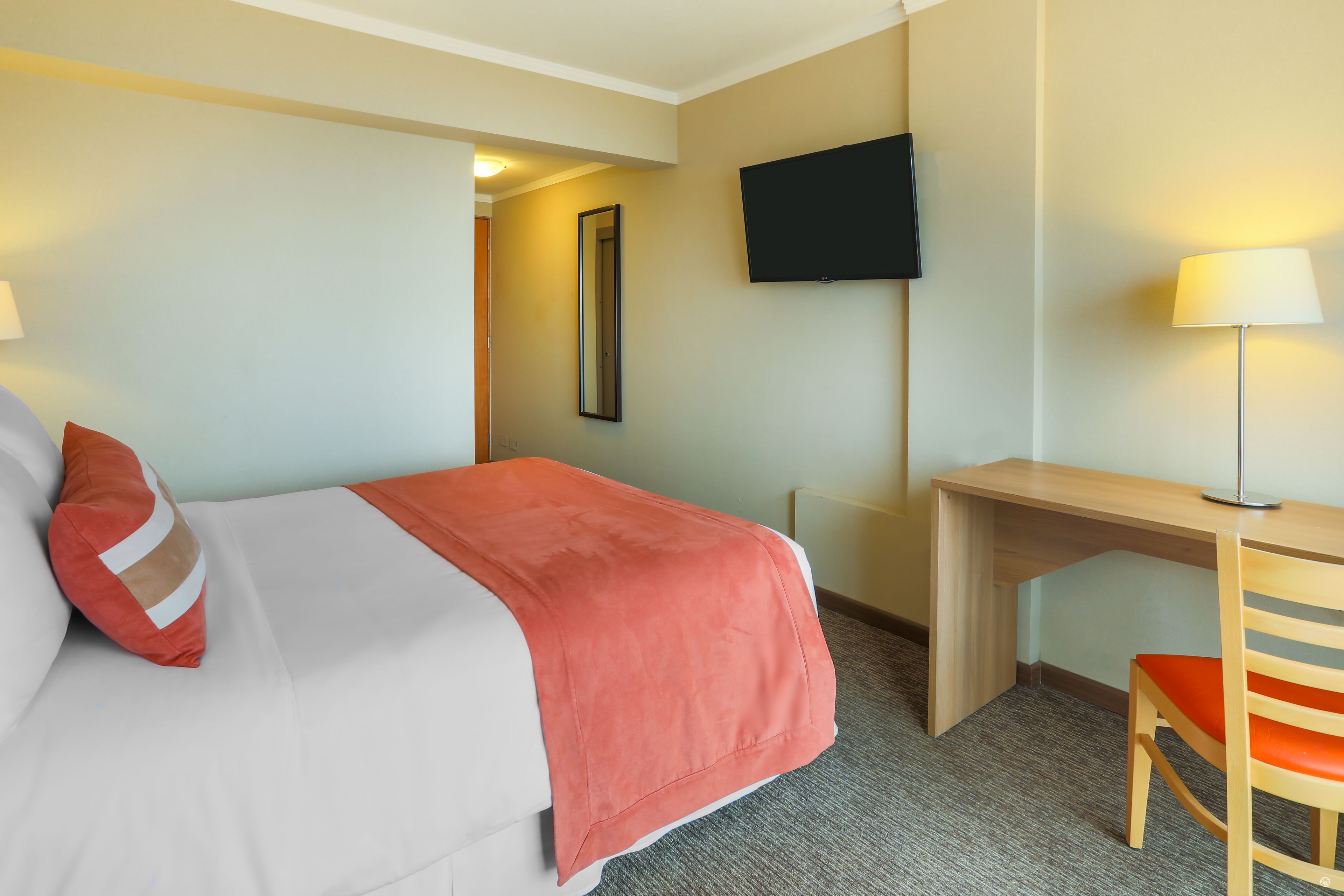 A hotel room with a bed, a desk, a chair, a TV, and a lamp.
