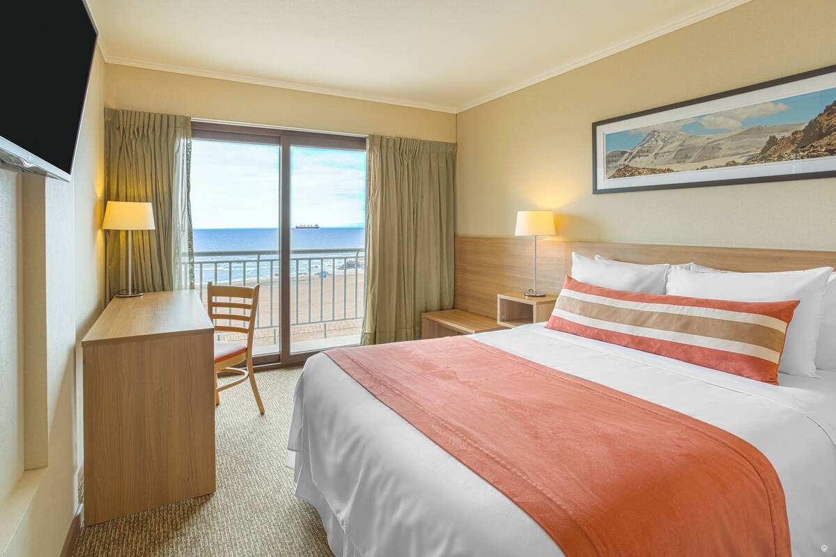 Een hotelkamer met een bed, een bureau, een stoel, een nachtkastje en uitzicht op zee door een schuifdeur.
