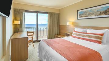 A hotel room with a bed, a desk, a chair, a nightstand, and a view of the sea through a sliding door.